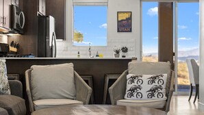 A modern kitchen with dark cabinetry, a stainless steel refrigerator, and a window with a scenic view. There are two chairs with cushions, one with a dinosaur print pillow.