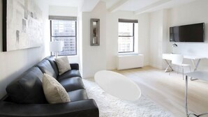 A modern living room with a black leather sofa, a white coffee table, a flat-screen TV, and large windows offering a city view.