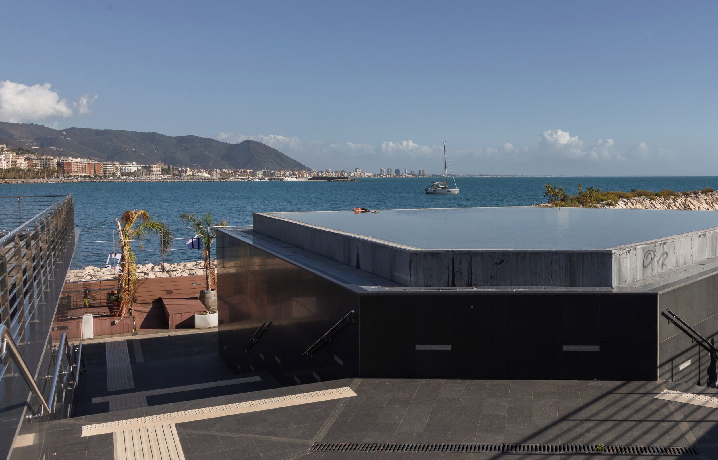 Una terrazza sul tetto con vista sul mare, un veliero e uno skyline sullo sfondo.
