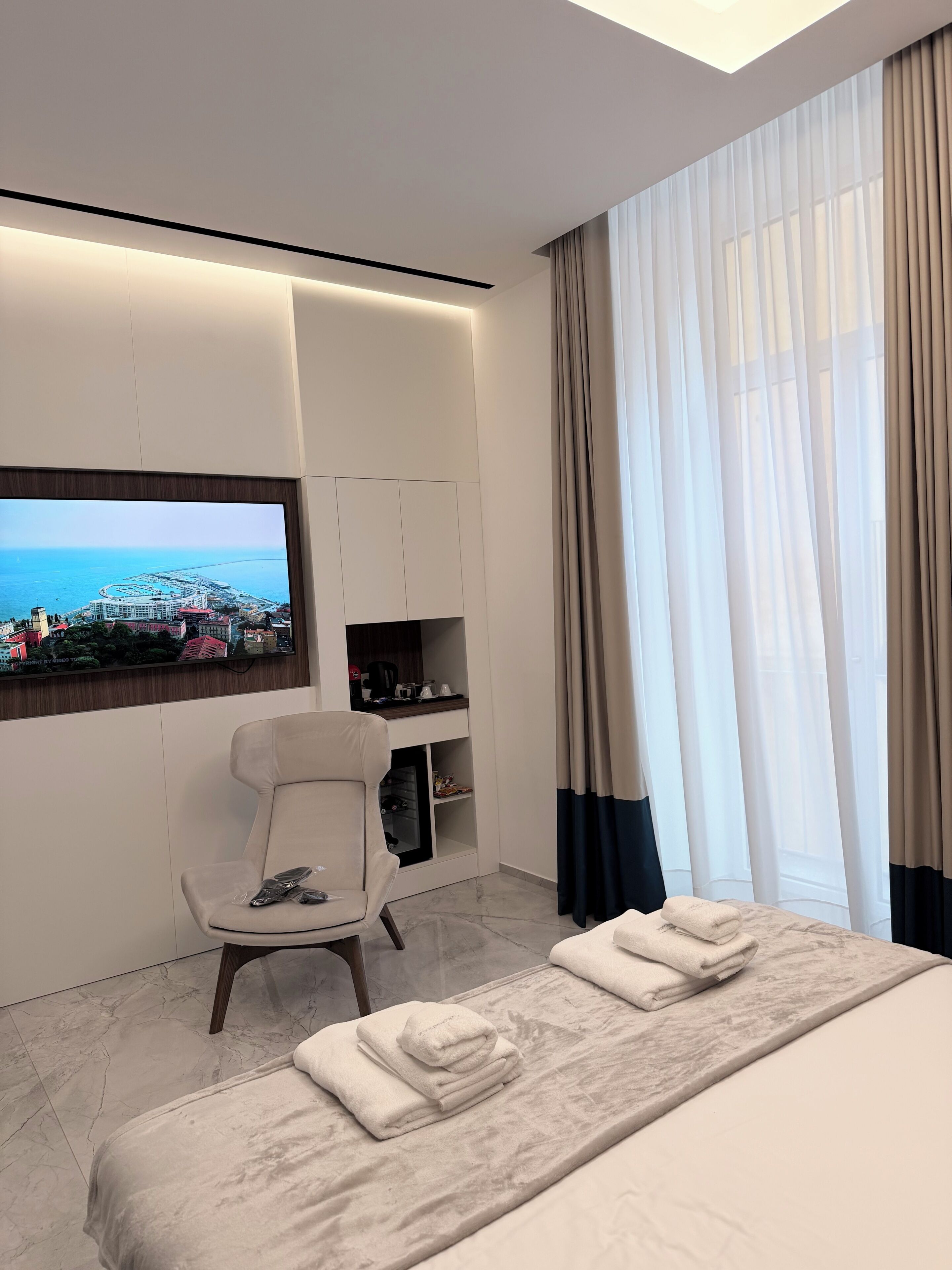 Ein modernes Hotelzimmer mit Bett, einer Sitzecke, einem Fernseher, der eine Stadtansicht zeigt, und Blick auf das Meer.