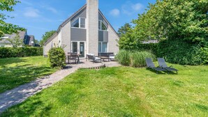 Een twee verdiepingen tellend huis met een schuin dak en schoorsteen, omgeven door een verzorgd gazon en een verhard pad dat naar de ingang leidt.