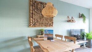 Un coin repas comprenant une table en bois, des chaises, une suspension en fibres tressées, une œuvre d’art murale, un téléviseur et des plantes en pot.