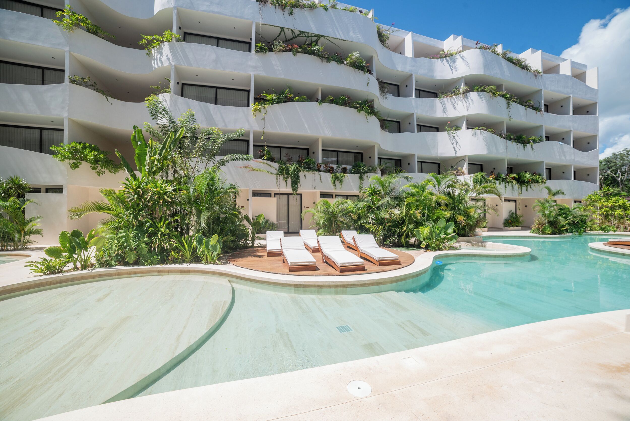 A multi-story hotel with balconies adorned with plants, a pool with lounge chairs, and tropical landscaping.