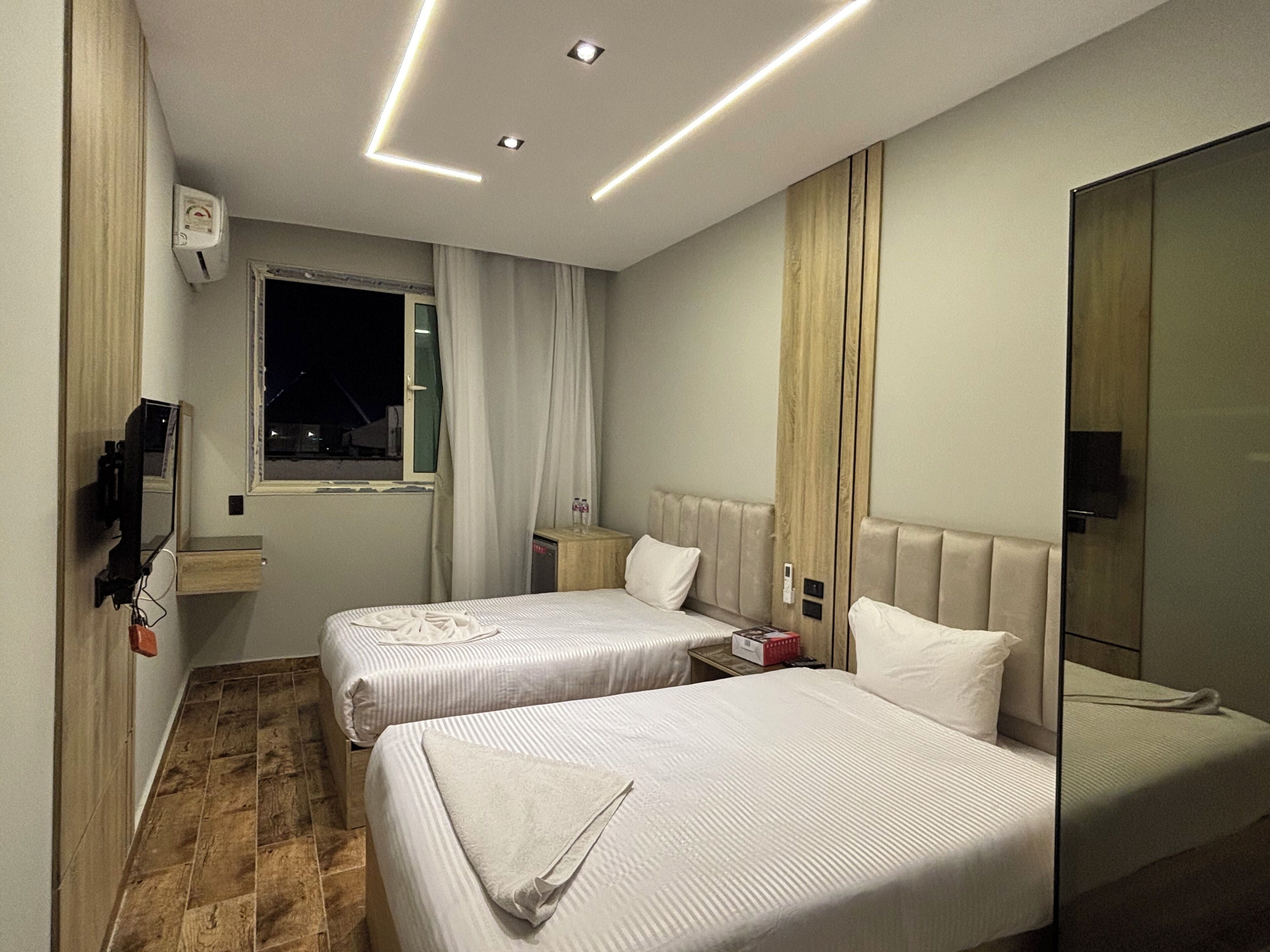 A hotel room with two beds, a window with curtains, a bedside table with a lamp, and a mirror.