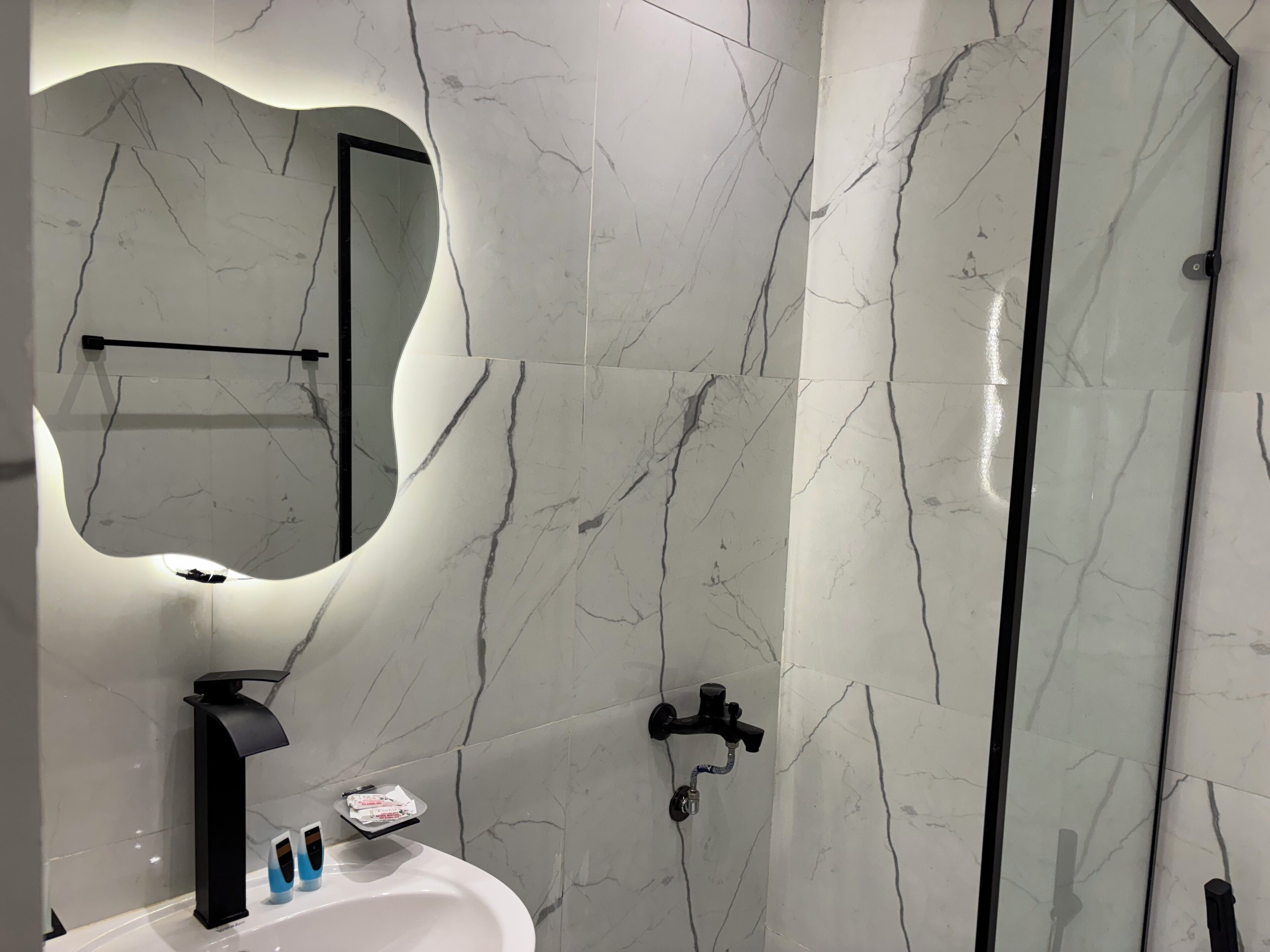 A modern bathroom with a white marble backsplash, a round mirror, a black faucet, and a glass shower enclosure.