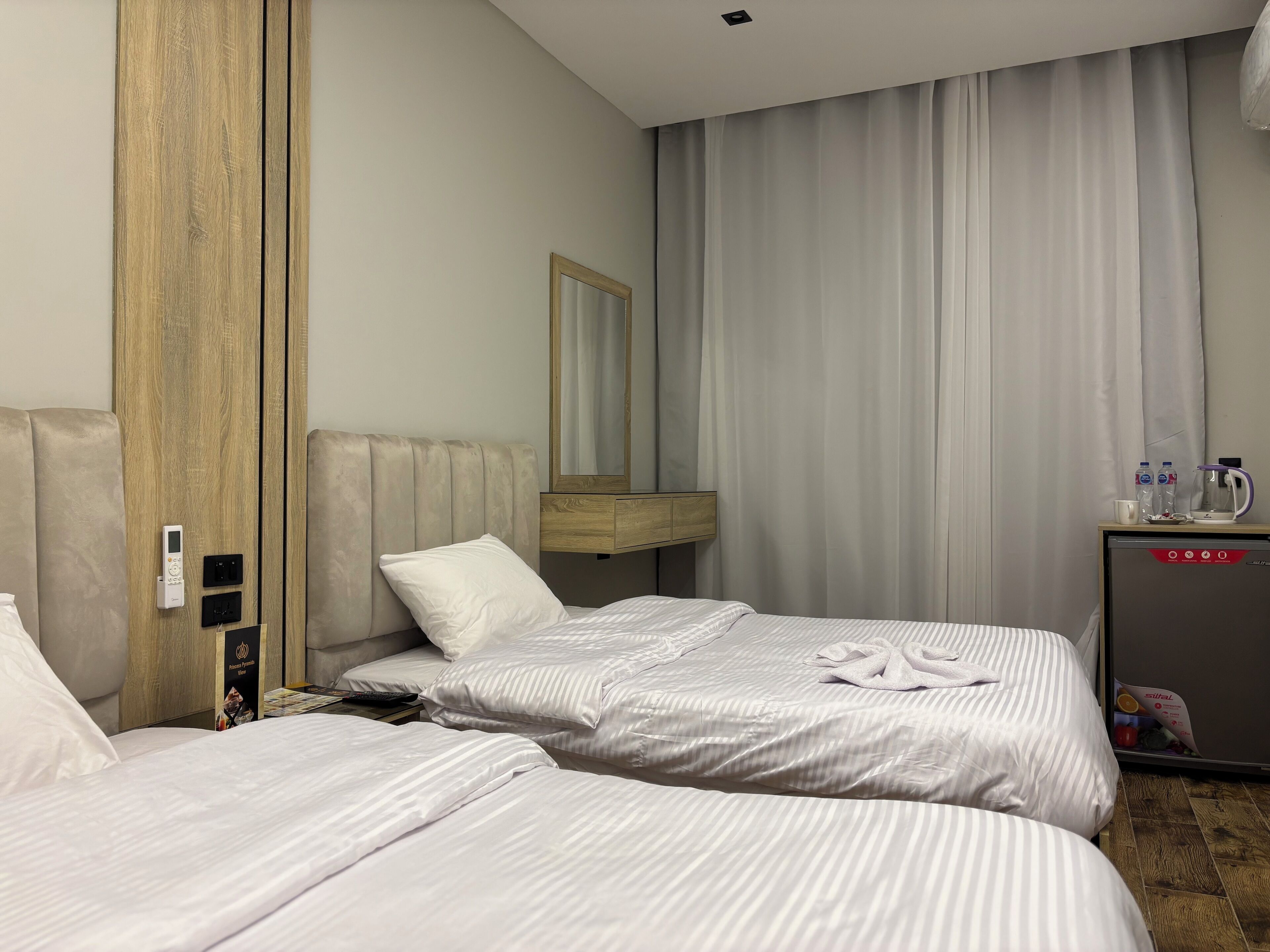 A hotel room with two beds, a wooden wardrobe, a bedside table with a mirror, a small cabinet, and a minibar.