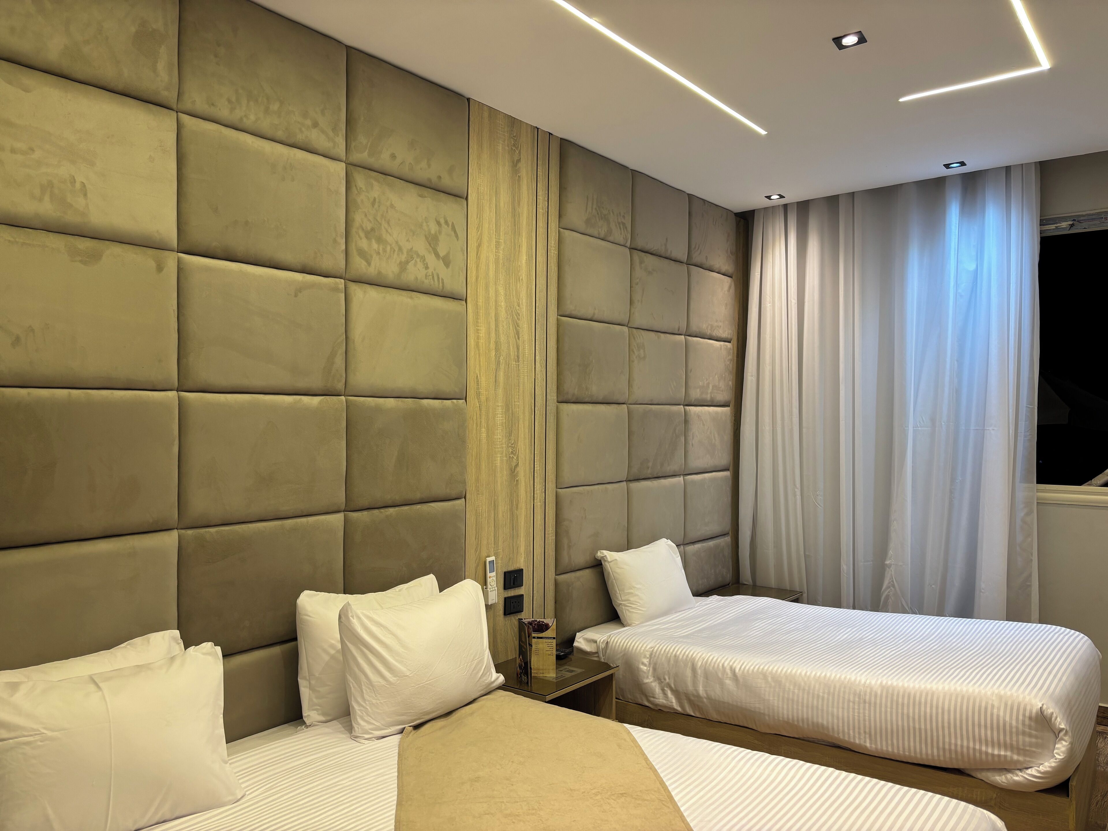 Une chambre d’hôtel moderne avec deux lits, une grande tête de lit rembourrée, une table de chevet et une fenêtre avec des rideaux.
