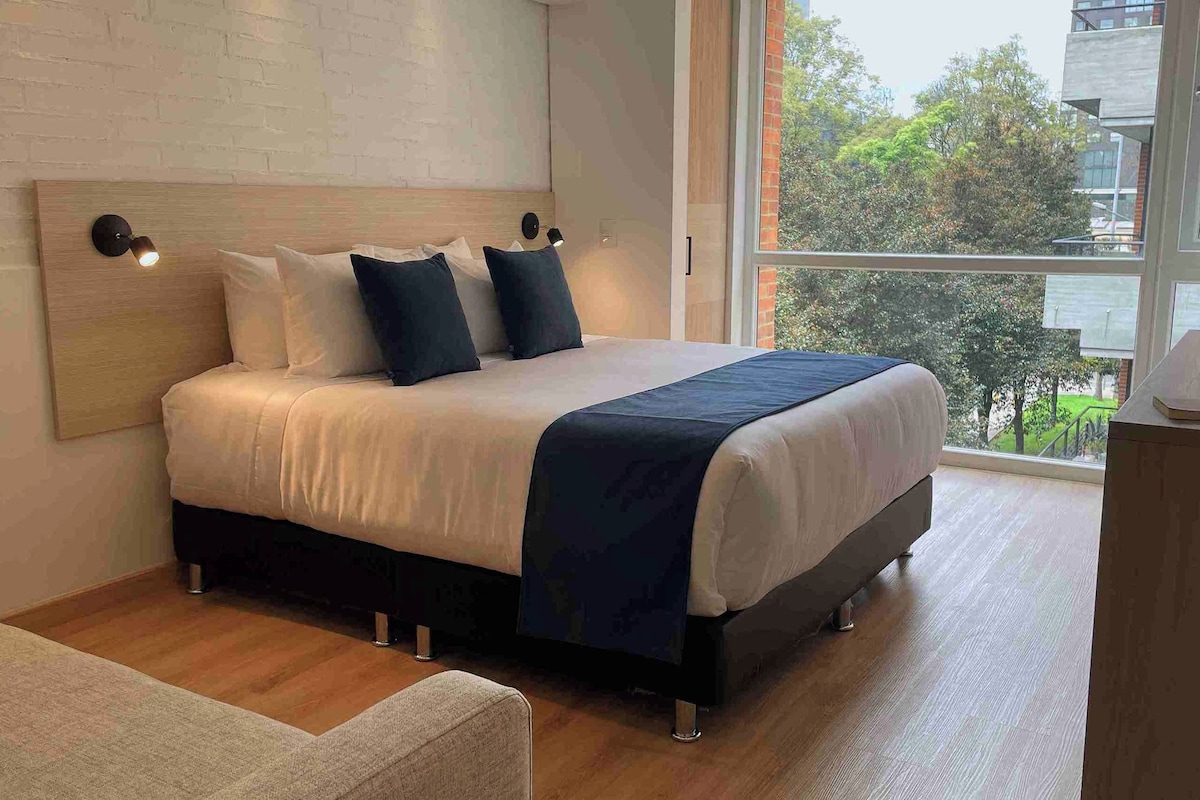 A modern bedroom with a bed, pillows, a bedside table, and a window with a view of buildings and trees.