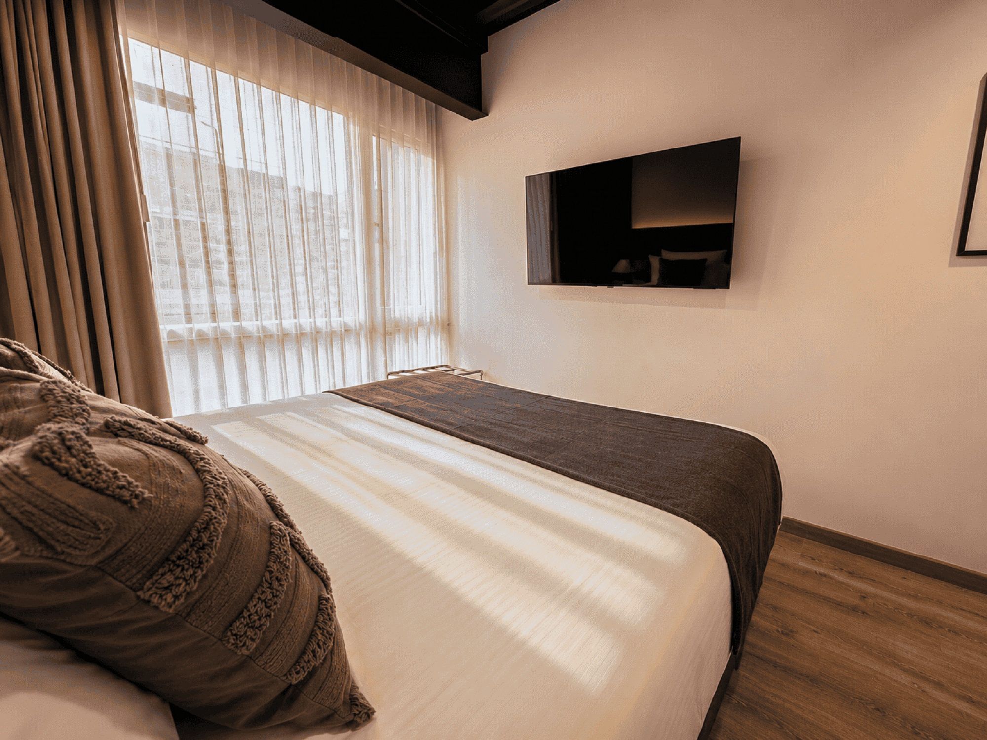 Un dormitorio con cama, ventana con cortinas, televisor montado y suelo de madera.