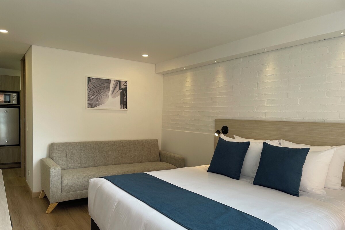 A hotel room with a bed, a sofa, a wall-mounted picture, and a refrigerator.