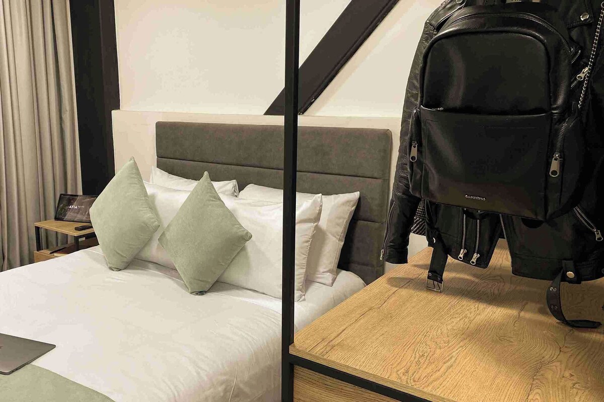 A hotel room with a bed, pillows, a laptop, a wooden nightstand, and a backpack.