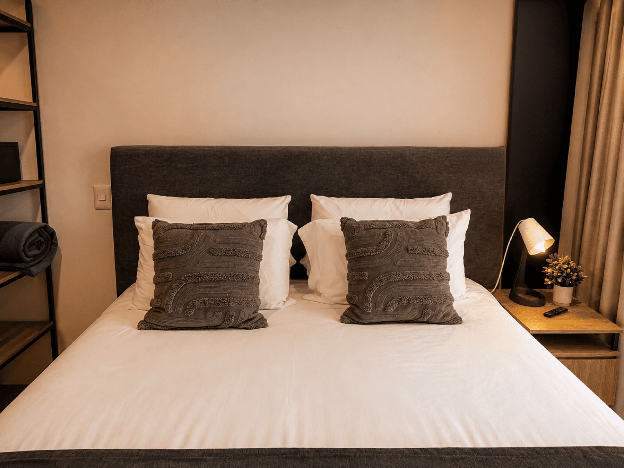 Una cama bien tendida con sábanas blancas y dos almohadas decorativas, una mesita de noche con una lámpara y una planta pequeña, y un estante con ropa doblada.