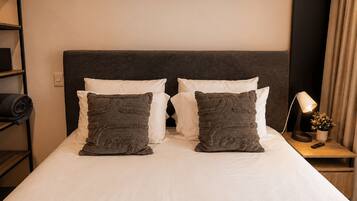 Una cama bien tendida con sábanas blancas y dos almohadas decorativas, una mesita de noche con una lámpara y una planta pequeña, y un estante con ropa doblada.