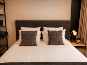 A neatly made bed with white linens and two decorative pillows, a bedside table with a lamp and a small plant, and a shelf with folded clothes. - Nova Iorque Suites by Avia Suites (Bogotá)