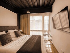 A hotel room with a large bed, a desk, and a chair. There is a window with curtains and a view of the outside. - Nova Iorque Suites by Avia Suites (Bogotá)