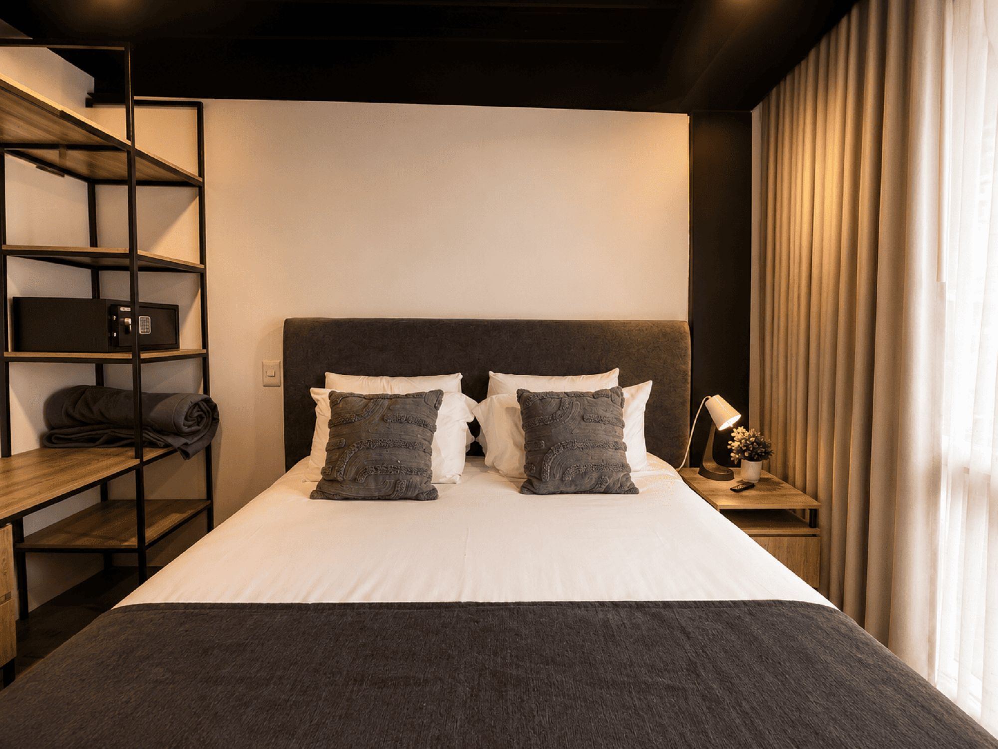 Una habitación de hotel con una cama, una mesita de noche con lámpara, un estante con caja fuerte y una ventana con cortinas.