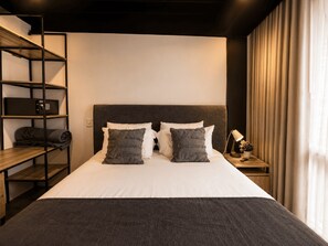 A hotel room with a bed, a nightstand with a lamp, a shelf with a safe, and a window with curtains. - Nova Iorque Suites by Avia Suites (Bogotá)