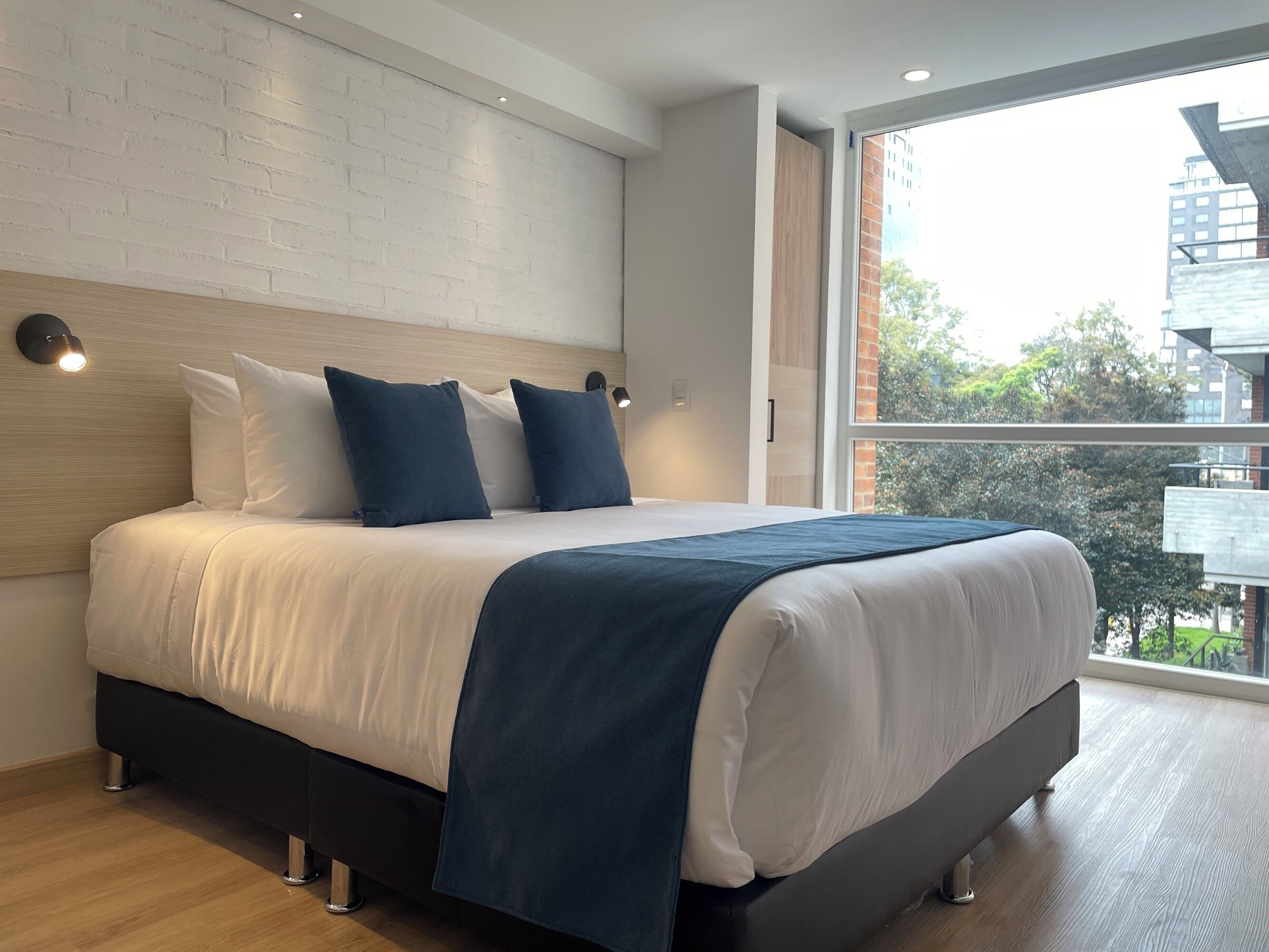 A modern hotel room with a large bed, a white brick wall, and a view of the city.