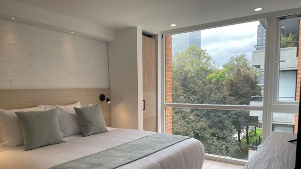 A modern bedroom with a large bed, a nightstand, a wall-mounted light, and a large window with a city view. - Nova Iorque Suites by Avia Suites (Bogotá)