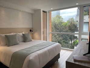 Un dormitorio moderno con una cama grande, una mesita de noche, una lámpara de pared y un ventanal con vistas a la ciudad.
