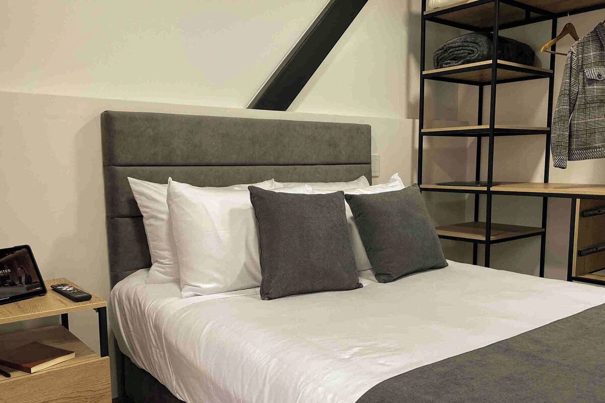 A neatly made bed with a grey headboard, a bedside table with a book and remote, and a clothing rack with a jacket.