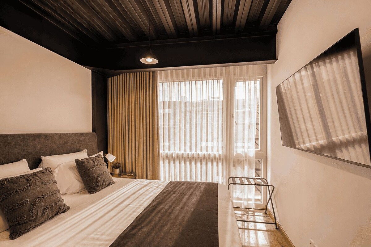 A hotel room with a large bed, a desk, and a chair. There is a window with curtains and a view of the outside.