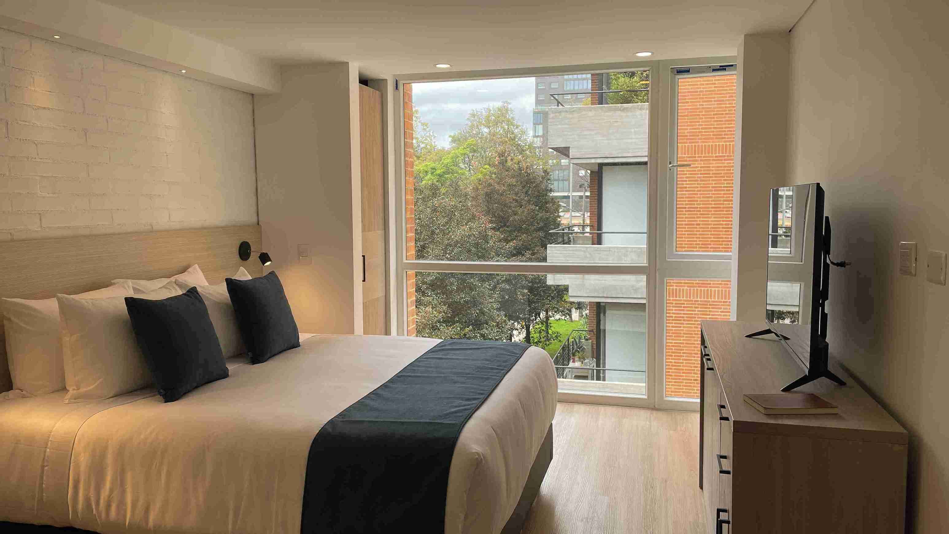 A modern bedroom with a large bed, a TV, and a balcony with a city view.
