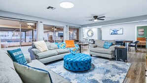 A spacious living room with a grey sofa, a blue ottoman, and a dining area with a table and chairs. - The Desert Oasis- Pool, Spa, Game Room, Basketball, Pickleball (Lake Havasu City)