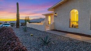 A stucco house with a paved driveway, a cactus, and a well-lit interior visible through an arched doorway. - The Desert Oasis- Pool, Spa, Game Room, Basketball, Pickleball (Lake Havasu City)