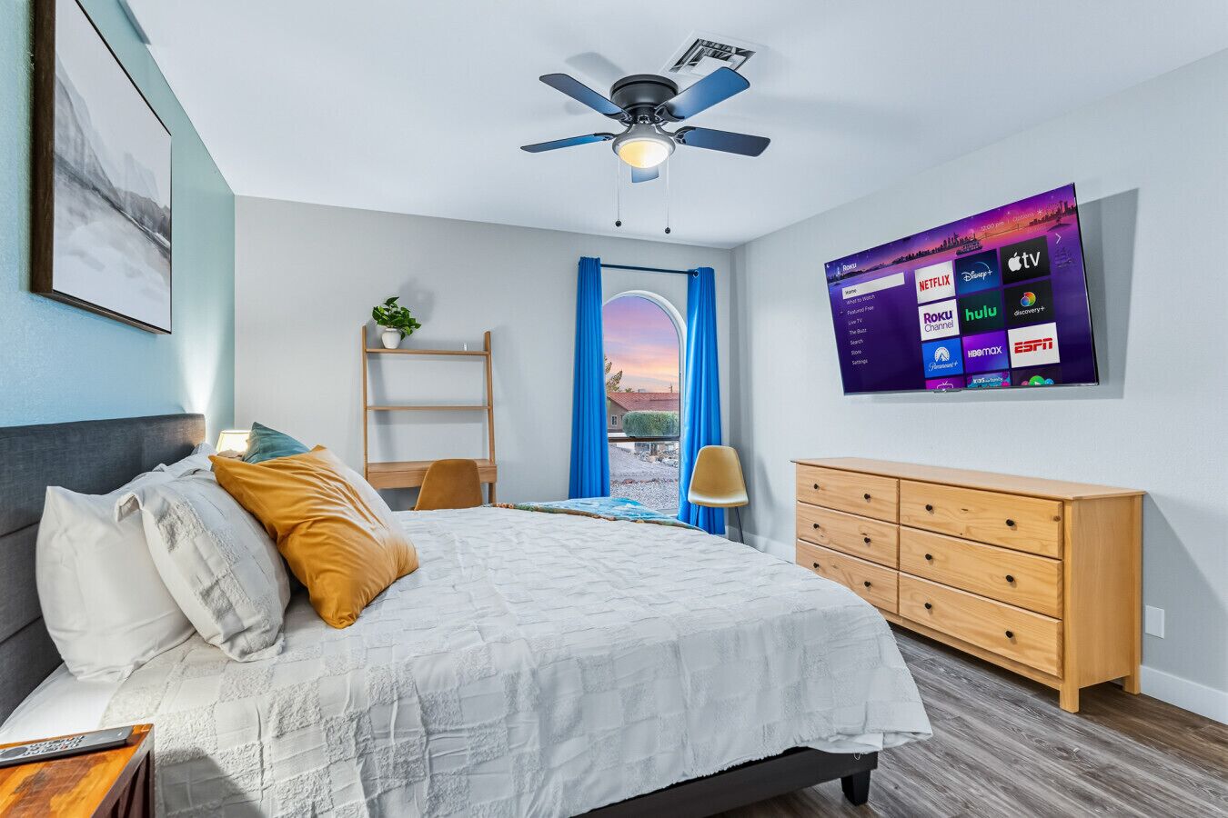 A modern bedroom with a bed, a desk, a dresser, a ceiling fan, and a TV displaying streaming services.