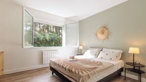 A bedroom with a large window, a bed with a blanket, two bedside tables with lamps, and a wall-mounted decorative item. - Villa Hortensia - Welkeys (Soorts-Hossegor)