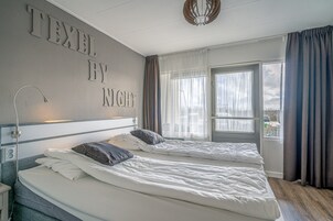 A hotel room with two beds, a large window with curtains, and a wall with the text "TEXTEL BY NIGHT".