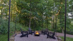 Un espacio al aire libre rodeado de bosque, con un hogar para fogata y sillas, luces colgantes y un sendero de grava.
