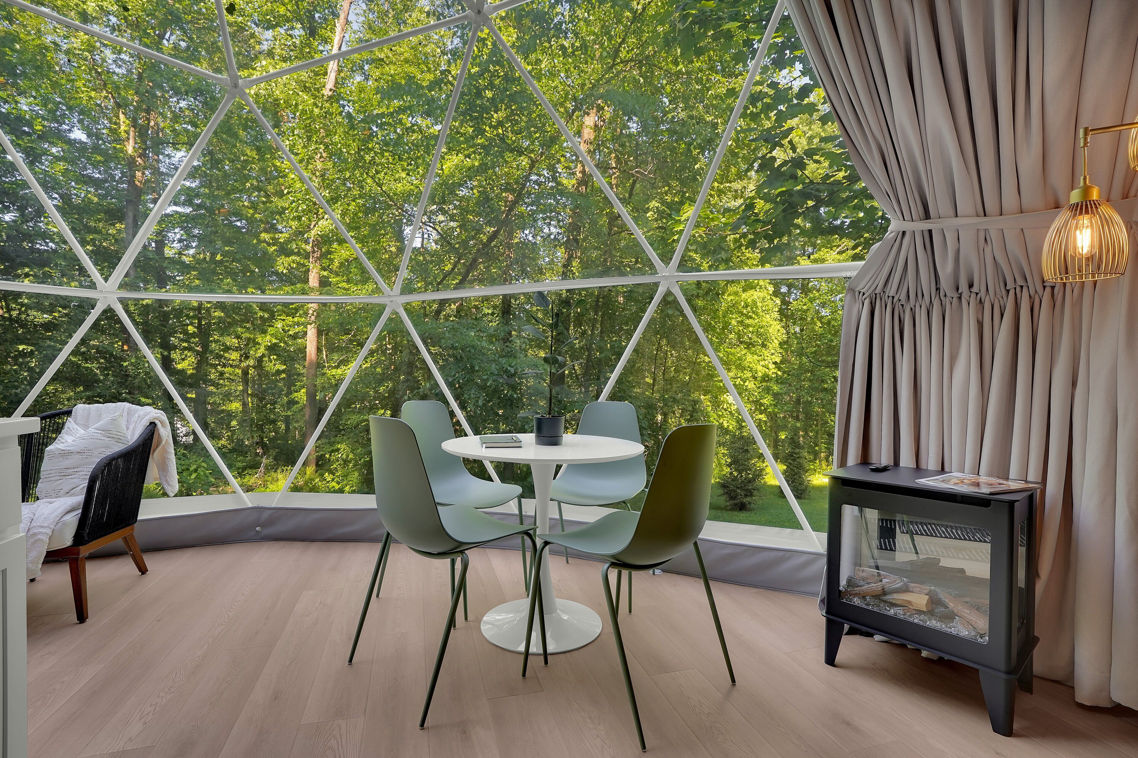 Ein Zimmer mit einer Glaskuppel, einem runden Tisch mit Stühlen, einem kleinen schwarzen Beistelltisch mit Lampe und Blick auf Bäume durch die Kuppel.