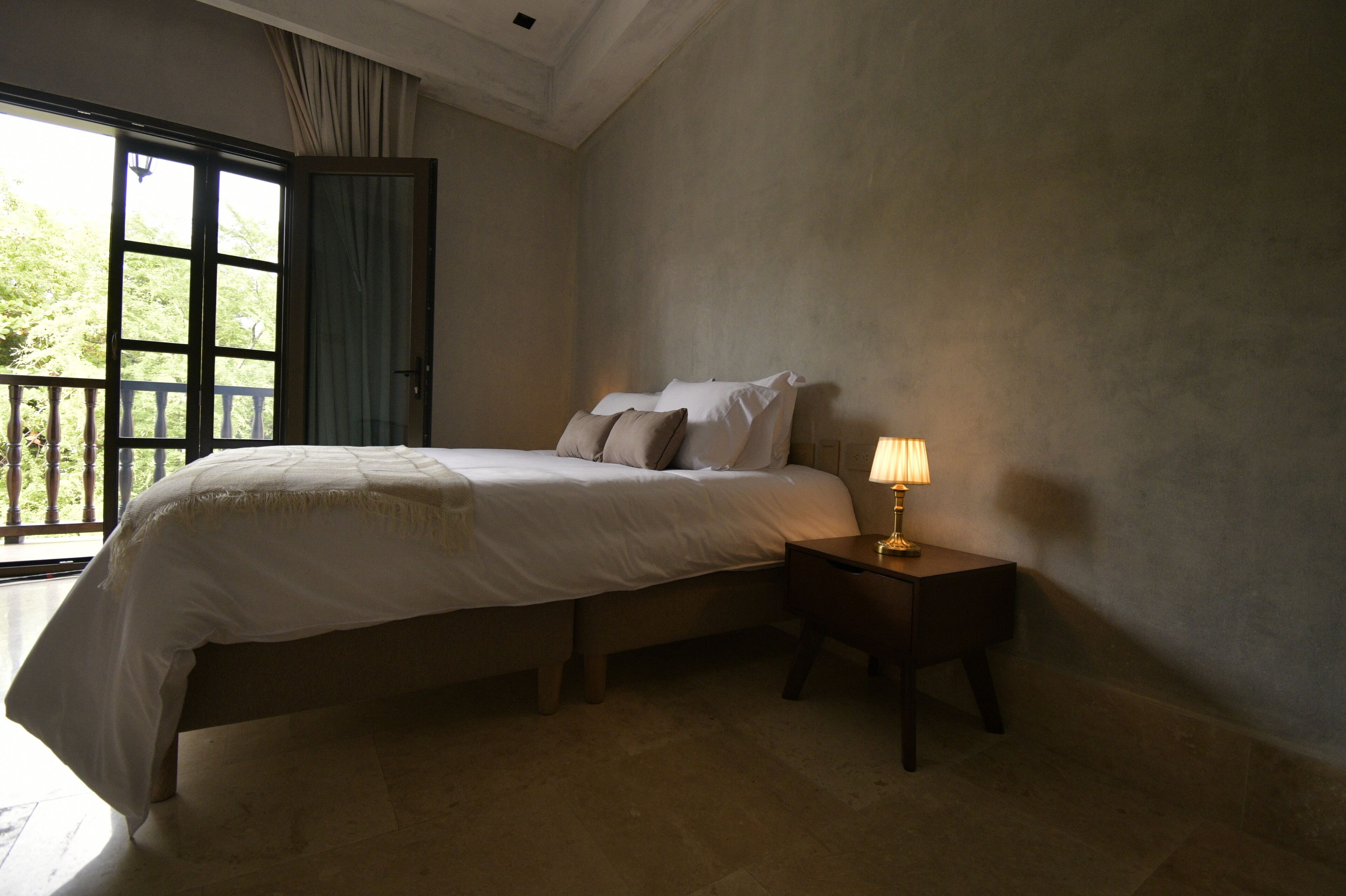 Una camera da letto con un letto, un comodino con una lampada, una finestra con tende e vista su del verde all'esterno.