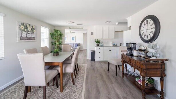 A modern kitchen with white cabinets, a wooden dining table with chairs, and a clock on the wall. - Gardenia Cottage | In The Heart of Pinecraft! (Sarasota)