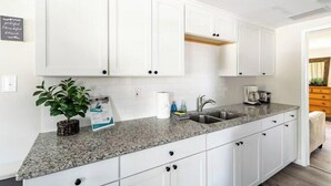 A kitchen with white cabinets, a granite countertop, a sink, and a potted plant. - Gardenia Cottage | In The Heart of Pinecraft! (Sarasota)
