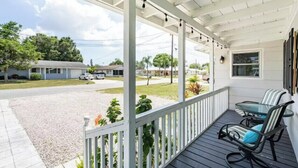 A covered porch with a table and chairs, a view of a street with houses, and a clear sky. - Gardenia Cottage | In The Heart of Pinecraft! (Sarasota)