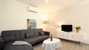 Un moderno salón con un sofá gris, una mesita redonda con una botella, un televisor de pantalla plana sobre un mueble blanco y un cuadro en la pared.
