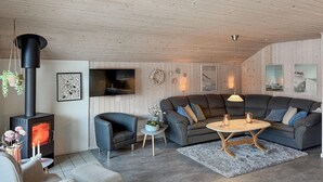 Een gezellige woonkamer met een houten plafond, een zwarte houtkachel, een grijze bank, een klein rond tafeltje en een aan de muur bevestigde televisie.