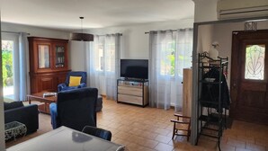 A living room with a TV, a blue armchair, a wooden cabinet, and a dining area with a table. - Gîte Les Alicantes - BAGES (BAGES)