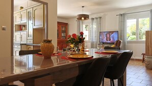 A dining area with a large table, chairs, a vase with flowers, and a television in the background. - Gîte Les Alicantes - BAGES (BAGES)