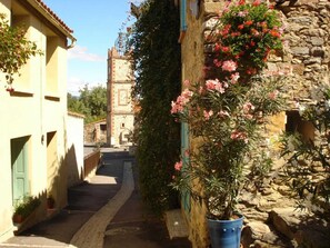 A narrow street with stone buildings, a bell tower, and a potted plant with pink flowers.