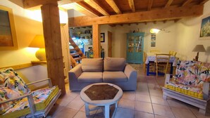 A cozy living room with a sofa, a small round table, and a colorful chair. There is a kitchen area in the background with cabinets and a dining table.