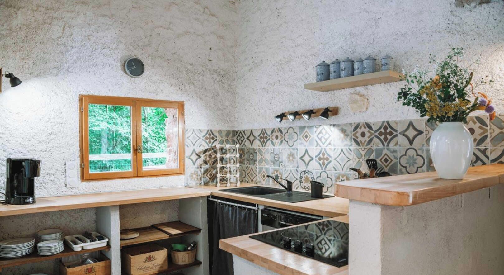 A kitchen with a wooden countertop, a sink, and a window with a view of greenery. There are shelves with kitchen items and a vase with flowers on the counter.