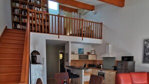 A dining area with a table, chairs, and a red sofa. A staircase leads to a loft with a bookshelf.