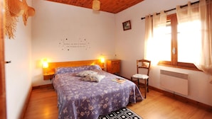 Une chambre avec un plafond en bois, un lit recouvert d’une couvre-lit à motifs floraux, une chaise, une table de chevet avec une lampe et un mur orné d’une citation décorative.