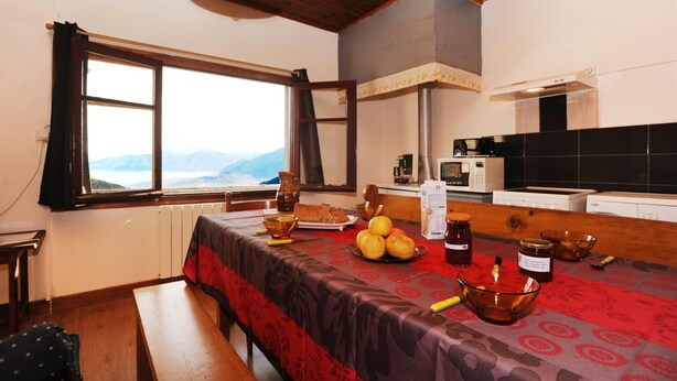 Un coin repas avec une table dressée pour deux personnes, une vue sur les montagnes par la fenêtre et une cuisine équipée d’appareils électroménagers modernes.