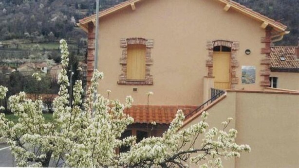 Ein zweistöckiges Gebäude mit flachem Dach und Schornstein. Zwei Fenster mit Fensterläden sind zu sehen, und im Vordergrund steht eine Straßenlaterne.
