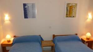 Ein Zimmer mit zwei Betten, jeweils mit blauer Bettdecke und einem hölzernen Kopfteil. Zwei Nachttische mit Lampen und ein kleiner gewobener Sessel stehen in der Mitte. Über den Betten hängen zwei gerahmte Kunstwerke.